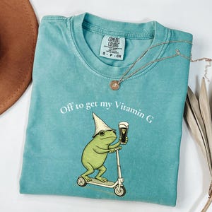May include: Teal Comfort Colors t-shirt with a frog graphic riding a scooter and holding a beer. The text "Off to get my Vitamin G" is printed on the shirt. A gold necklace with a pendant is also visible.