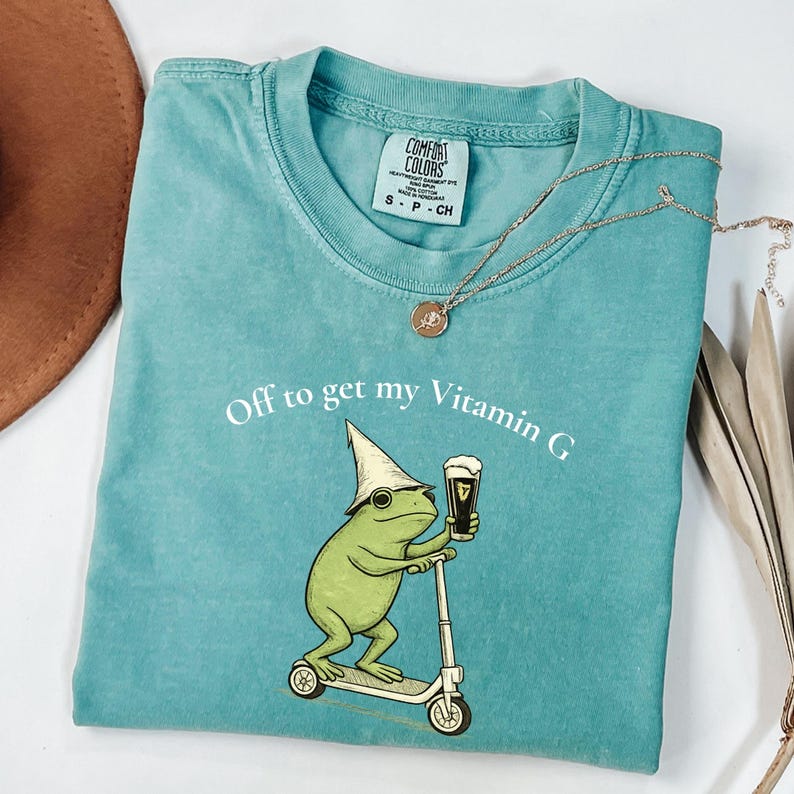 May include: Teal Comfort Colors t-shirt with a frog graphic riding a scooter and holding a beer. The text "Off to get my Vitamin G" is printed on the shirt. A gold necklace with a pendant is also visible.
