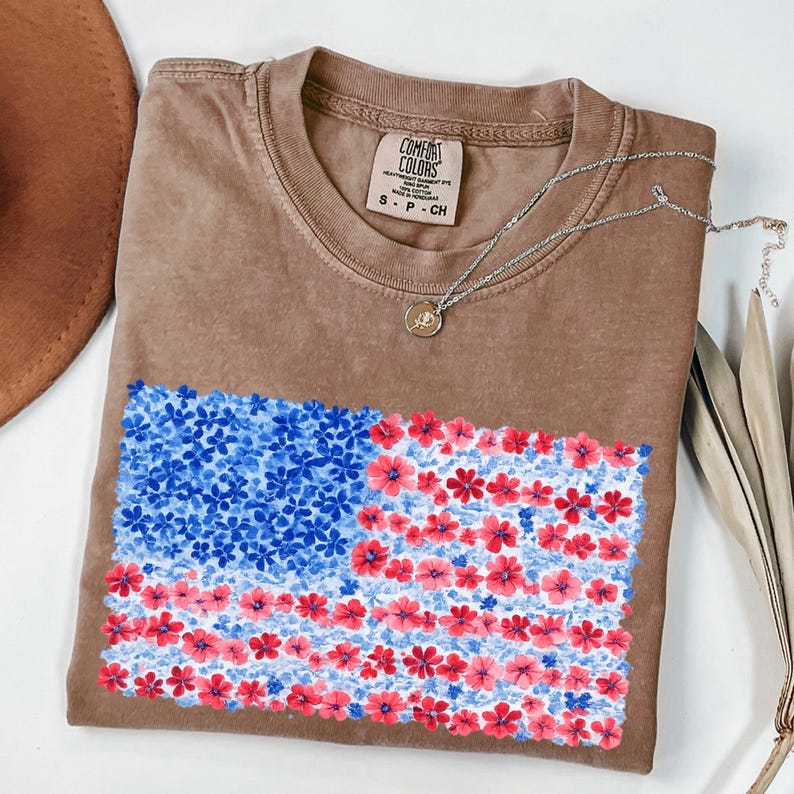 Puede incluir: Una camiseta marr&oacute;n con un dise&ntilde;o floral de la bandera estadounidense. La bandera presenta flores azules en la esquina superior izquierda y flores rojas para las rayas. Un collar plateado con un peque&ntilde;o colgante est&aacute; sobre la camisa.