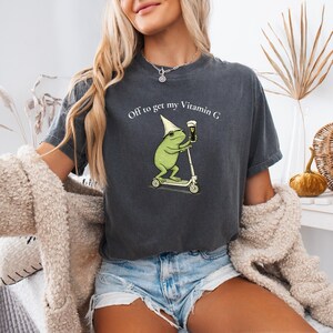 May include: Dark gray t-shirt featuring a cartoon frog riding a scooter and holding a beer. The frog wears a pointed hat and the shirt reads "Off to get my Vitamin G". The shirt is a casual, unisex style.