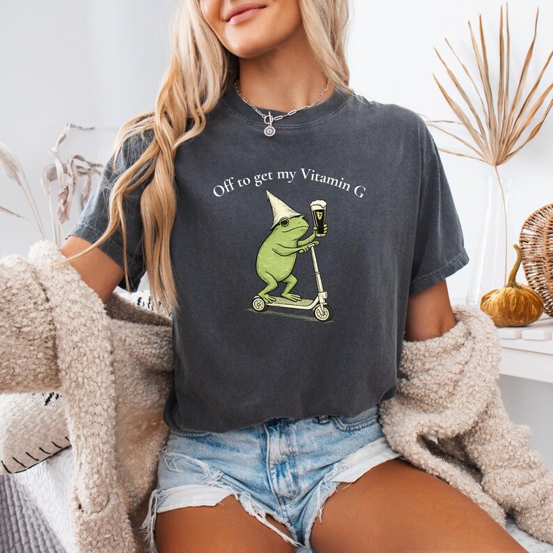 May include: Dark gray t-shirt featuring a cartoon frog riding a scooter and holding a beer. The frog wears a pointed hat and the shirt reads "Off to get my Vitamin G". The shirt is a casual, unisex style.