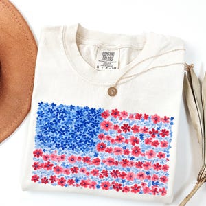 Puede incluir: Camiseta color crema con un dise&ntilde;o floral de la bandera estadounidense. La bandera est&aacute; compuesta por flores azules y rojas. Un collar dorado con un colgante descansa sobre la camiseta doblada sobre una superficie blanca.