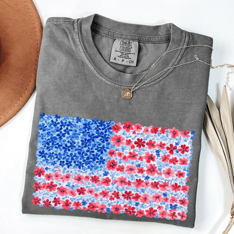 Puede incluir: Una camiseta gris Comfort Colors con un dise&ntilde;o floral de la bandera estadounidense en rojo y azul. La camiseta tiene cuello redondo y mangas cortas. Un collar dorado con un colgante est&aacute; drapeado sobre la camiseta.