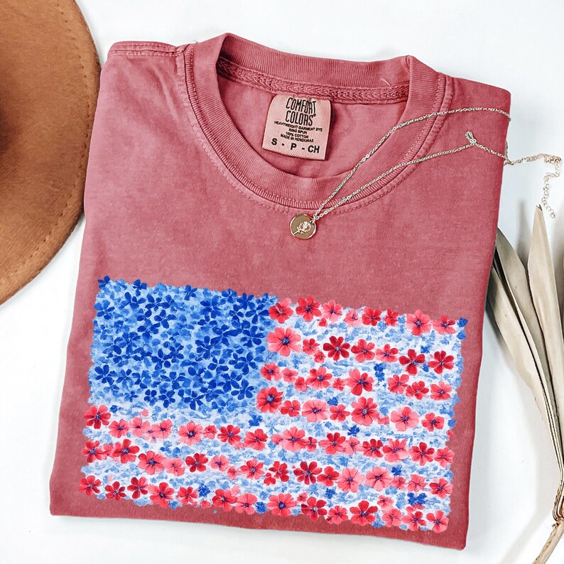 Puede incluir: Una camiseta Comfort Colors granate con un dise&ntilde;o floral de la bandera estadounidense. Las estrellas de la bandera son flores azules y las rayas flores rojas. Un collar con colgante dorado descansa sobre la camisa.
