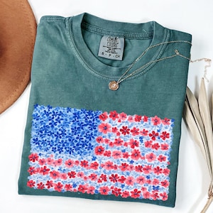 Puede incluir: Una camiseta Comfort Colors verde azulado con un dise&ntilde;o floral de la bandera estadounidense en rojo y azul. Un collar dorado con un peque&ntilde;o colgante descansa sobre la camisa. Un sombrero marr&oacute;n y tallos de plantas secas est&aacute;n en el fondo.