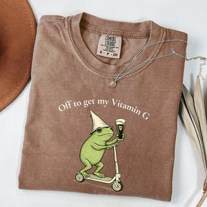 May include: Brown t-shirt with the text "Off to get my Vitamin G" and a graphic of a green frog on a scooter holding a beer. The shirt has a crew neck and short sleeves. A silver necklace with a small pendant is also visible.