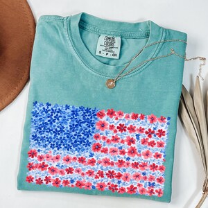 Puede incluir: Una camiseta Comfort Colors turquesa con un dise&ntilde;o floral de la bandera americana en rojo y azul. Un collar dorado con un colgante descansa sobre la camiseta. La camiseta est&aacute; doblada y expuesta sobre una superficie blanca.
