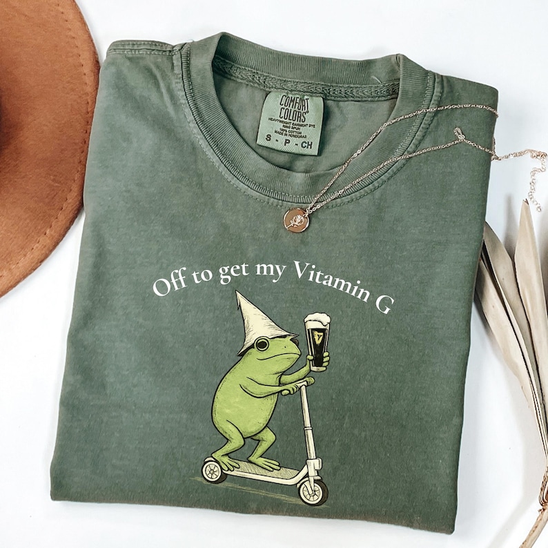 May include: Green Comfort Colors t-shirt with the phrase "Off to get my Vitamin G." The graphic features a frog on a scooter holding a beer. A gold necklace and a brown hat are also visible.