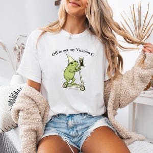 May include: White t-shirt with a whimsical graphic of a green frog riding a scooter and holding a pint of beer. The text "Off to get my Vitamin G" is printed above the frog. The shirt is a casual, everyday wear item.