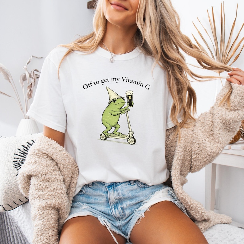 May include: White t-shirt with a whimsical graphic of a green frog riding a scooter and holding a pint of beer. The text "Off to get my Vitamin G" is printed above the frog. The shirt is a casual, everyday wear item.