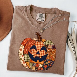 May include: A brown short-sleeve t-shirt featuring a large pumpkin graphic made of various patchwork patterns including autumn leaves, plaid, and floral prints. The pumpkin has a dark navy blue jack-o'-lantern face. The shirt tag reads Comfort Colors.