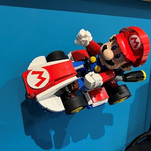 May include: A Lego model of Mario in a red kart with the number 3 on the side. The kart has black tires with yellow rims. Mario is wearing his signature red hat and overalls.
