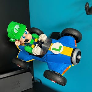 Puede incluir: Un modelo Lego de Luigi en un kart azul y amarillo con ruedas negras. Luigi lleva un gorro verde con una "L" blanca. El kart tiene una "L" blanca en el lateral.