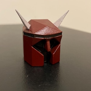 Può includere: Un piccolo elmo rosso e nero con due corna argentate. L'elmo ha un design geometrico e sembra essere stampato in 3D. L'elmo è appoggiato su una superficie scura.