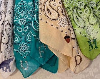May include: A collection of colorful bandanas with embroidered designs. The bandanas feature paisley patterns, letters, and other motifs like an elephant, horseshoe, acorn, and clover. Colors include white, teal, tan, and green.