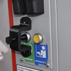 Puede incluir: Primer plano de un panel de control de una gasolinera. El panel es gris con una manija negra y un botón amarillo. Hay dos pegatinas con fondos azules y verdes. El panel está montado sobre una superficie gris.