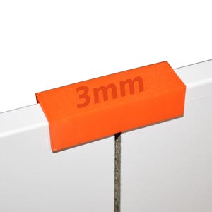 May include: A bright orange L-shaped plastic clip or guide piece featuring the text 3mm printed on the top surface. The object is positioned over the edge of a white panel, showing a clear 3mm measurement for alignment or spacing purposes.