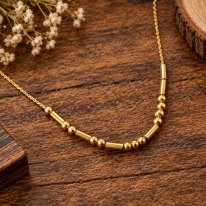 May include: A gold-toned necklace featuring a delicate chain adorned with a sequence of small, polished spherical beads and elongated cylindrical tube beads arranged in a repeating pattern along the center section of the jewelry piece.