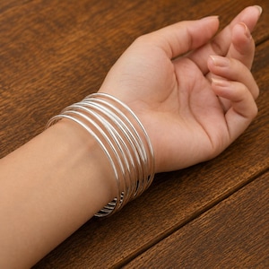Peut inclure: Un ensemble de sept joncs fins en métal argenté poli portés ensemble au poignet. Les bracelets présentent un profil lisse et arrondi avec une finition métallique réfléchissante, créant un aspect empilé qui entoure le bas du bras.