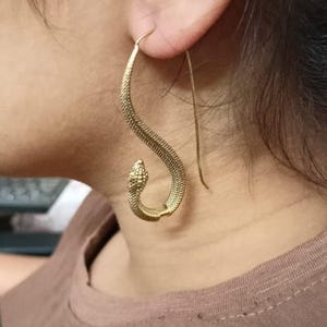 May include: A gold-toned earring shaped like a curved snake with a textured, scale-like surface. The earring features a long, thin wire hook that passes through the earlobe, allowing the snake body to hang vertically with the head pointing downward.