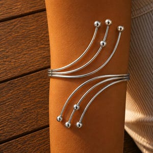 May include: A silver-toned arm cuff featuring a multi-strand design that curves around the arm. The piece consists of six thin, polished metal wires that fan out, each ending in a small, smooth spherical bead at the terminal points.