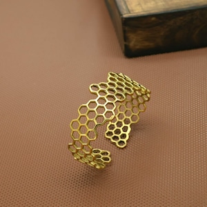 Può includere: Un bracciale a fascia color oro caratterizzato da un motivo a nido d'ape traforato. Il design è composto da forme esagonali interconnesse che creano una fascia ampia e strutturata con apertura sul retro e finitura metallica.
