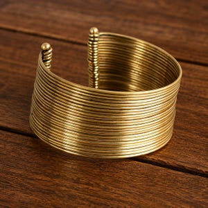 Peut inclure: Un large bracelet manchette composé de multiples fils métalliques fins et empilés de couleur dorée. Le design présente une série continue de bandes circulaires horizontales formant une structure rigide ouverte avec des embouts sphériques.