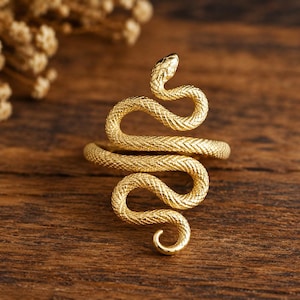 Könnte beinhalten: Ein goldfarbener Ring in Form einer gewundenen Schlange mit einer texturierten, schuppenartigen Oberfläche. Der Körper der Schlange bildet den Ring, wobei der Kopf oben positioniert ist und der Schwanz sich anmutig am unteren Ende krümmt.