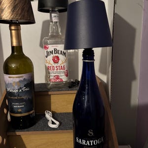 May include: Three bottle lamps featuring glass bases from Ménage à Trois, Jim Beam Red Stag, and Saratoga Still. Each bottle is topped with a small fabric lampshade in bronze, black, or navy blue. White charging cables are visible near the bases.
