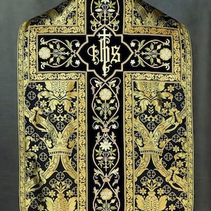 May include: Black and gold liturgical vestment with intricate floral and cross embroidery. The vestment features a central cross design with the letters IHS, surrounded by detailed gold patterns and floral motifs. The overall design is symmetrical and ornate.