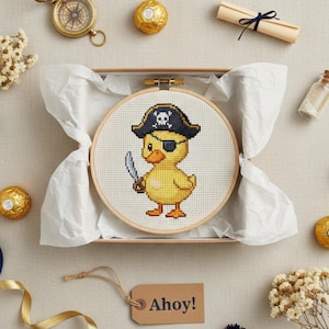 May include: A cross-stitch embroidery of a yellow duck wearing a pirate hat and eye patch, holding a sword. The embroidery is in a wooden hoop, presented in a gift box with a tag that says "Ahoy!". The scene includes a compass, scroll, and chocolates.