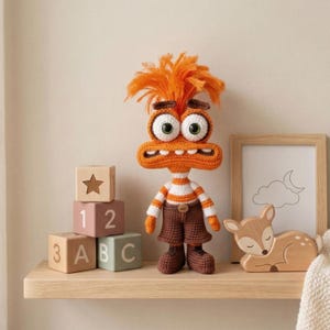 May include: A handmade orange and brown crochet monster doll with large eyes, a striped shirt, and brown shorts. The doll is standing on a wooden shelf next to wooden blocks and a wooden deer figurine. The doll has orange hair.