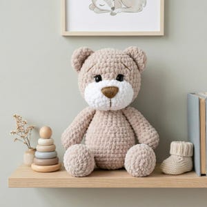 May include: A handmade, beige crochet teddy bear with a white muzzle and brown nose sits on a wooden shelf. The bear is surrounded by a wooden stacking toy, a small vase with dried flowers, and a knitted baby bootie. A framed picture of a sleeping deer is in the background.