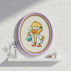 May include: Cross-stitch art of a yellow duck wearing safety glasses and a lab coat, holding a flask with green liquid. The artwork is framed in a light purple oval frame with a gold inner border. The duck is a cartoon character.