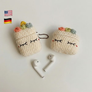 May include: Two off-white, crocheted AirPod cases with closed-eye designs and colorful floral accents. The cases are next to a pair of white AirPods. A small chain is attached to one case.