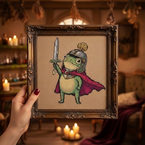 May include: Cross-stitch art of a frog warrior in a helmet and cape, holding a sword aloft. The framed artwork features a detailed design with green, red, and silver accents. The frame is ornate, with a gold finish. The artwork is displayed in a room with candles.