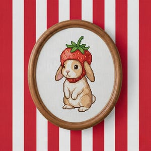 May include: A framed cross-stitch of a bunny wearing a strawberry hat and collar. The bunny is tan and white, with pink ears and nose. The strawberry hat is red with green leaves. The frame is round and made of wood. The background is red and white striped.