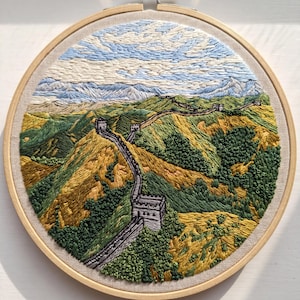 Op de afbeelding: Handgemaakte borduurkunst van de Chinese Muur. Het kunstwerk toont een gedetailleerd landschap met glooiende heuvels, een kronkelende muur en een blauwe lucht. De borduursel is geplaatst in een ronde houten hoepel.