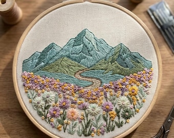 May include: Embroidered mountain landscape in a wooden hoop. The artwork features teal and green mountains, a path, and a field of flowers in yellow, purple, and white. The embroidery is on cream fabric. Includes embroidery thread, needles, and scissors.
