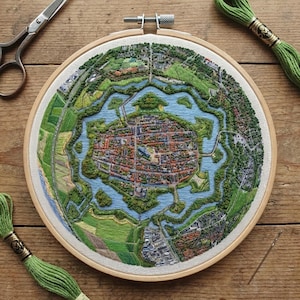 Op de afbeelding: Een gedetailleerde borduurwerk van een stadskaart in een houten ring. Het kunstwerk toont een centraal stedelijk gebied omgeven door water en groene landschappen, met ingewikkelde steken in verschillende kleuren. Schaar, draad en een naald zijn zichtbaar.