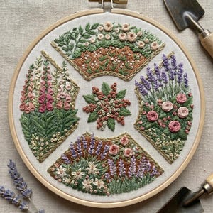 Può includere: Design floreale ricamato in un cerchio di legno. L'opera presenta vari fiori nei toni del rosa, viola, verde e marrone. Due piccole cazzuole da giardino e rametti di lavanda sono posizionati attorno al cerchio.