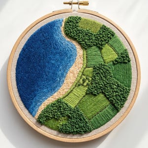 Puede incluir: Bordado de paisaje en un aro de madera. La obra representa una escena costera con un océano azul, una playa de arena y campos verdes. El bordado utiliza varios tonos de azul, verde y beige para crear profundidad y textura.