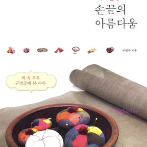 Op de afbeelding: Een boekomslag met Koreaanse tekst en kleurrijke patchwork speldenkussens in een houten kom. De titel van het boek is in het Koreaans, met extra tekst en illustraties. Een opgerold stuk stof staat op de achtergrond.