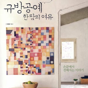 May include: A book cover with Korean text and a colorful geometric artwork. The artwork is a patchwork of squares in various colors. A wooden bowl with green fruit sits on a wooden table. The book title is "Gyubang Gongye".