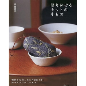 May include: A book cover with Japanese text and a small, embroidered pouch. The pouch is dark blue with green leaf designs and sits in a white bowl. Other white bowls and a cup are in the background.