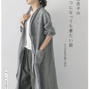 Pode incluir: Uma mulher vestindo um casaco longo de linho cinza com bolsos, combinado com calças cinza e uma blusa branca. Texto japonês e o design da capa de um livro são visíveis.