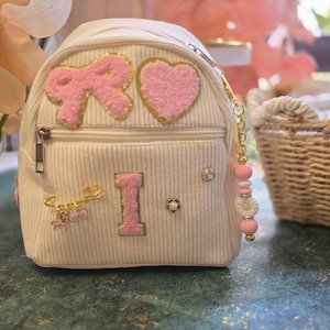 May include: A white corduroy mini backpack featuring a pink fuzzy bow patch, a pink fuzzy heart patch, and a pink fuzzy letter I patch. The bag includes a gold safety pin with small flower charms, a pearl-like stud, and a beaded keychain.