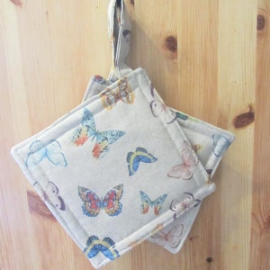 May include: Two beige pot holders with a butterfly pattern. The pot holders have a loop for hanging.