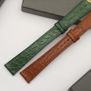 Könnte beinhalten: Zwei Uhrenarmbänder mit einer strukturierten Alligator-Optik. Ein Armband ist dunkelgrün und das andere mittelbraun. Beide Bänder verfügen über eine Reihe von gestanzten Löchern zur Anpassung, eine feste Schlaufe und eine bewegliche Schlaufe.