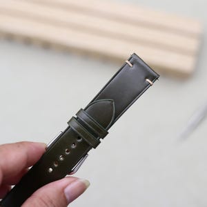 May include: A dark olive green watch strap featuring a smooth finish, two matching leather keepers, and light-colored stitching near the lug end. The strap includes a series of punched holes for adjustment and a silver-toned metal buckle component.